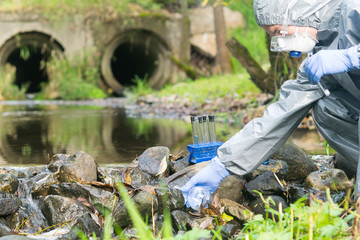 a man in special protective clothing and a mask draws water from a river into a flask after discharges into the river, checking the river for environmental friendliness