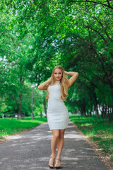 Portrait of a charming blond woman wearing beautiful white dress standing on the road under trees.