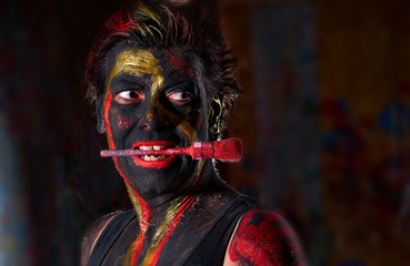 portrait of an attractive European (Caucasian) lady with black, golden and red body painting against dark background - she has a round red brush across her face and looks lasivously to the right side - obrazy, fototapety, plakaty