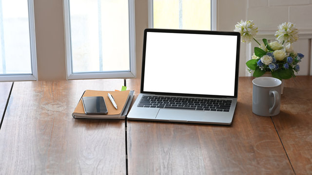 Photo of white blank screen laptop, coffee cup, notebook, pen and potted plant putting together on modern wooden table with light through the window as background.