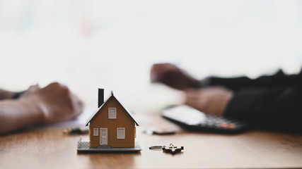 Photo of house model and key putting on the modern wooden desk with hand of property agency and customer as background.