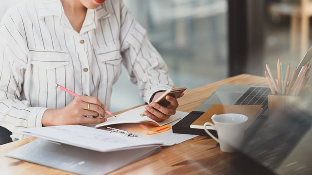 Cropped shot of young beautiful woman in striped shirt holding smartphone in hand and writing/taking note white sitting at the modern wooden desk with modern office as background.
