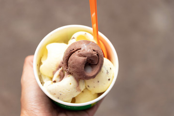 Hand holding take away cup of ice cream with street background. Happy walking and eating in hot weather with street dessert stall in Thailand.
