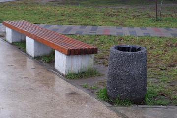 Stone black trash can with white splashes stands near brown bench on white blocks-legs near concrete walking area wet from rain, behind the bench can see lawns with low green grass and mosaic sidewalk