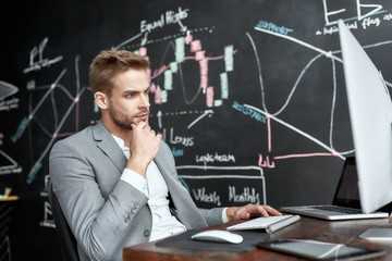 For the real difference. Young trader sitting by desk in front of computer monitor while working in the office. Blackboard full of charts and data analyses in background.