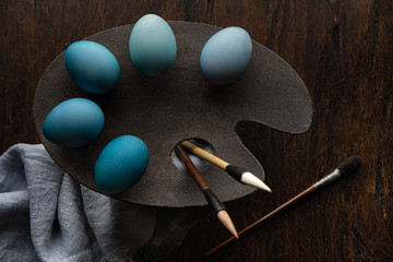 Paint brushes and colored eggs in different shades of blue on the artist's palette. Dark photo. Dark and light. Wooden background. Blue gradient