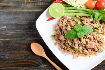 Thai food, spicy minced pork salad (Larb Moo) on white plate with spoon on wooden background, top view