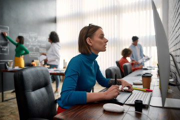 Graphic designer at work. Side view of young woman using digital graphics tablet while sitting at...