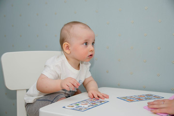 A small child sits at a white table and plays in a children's lotto. The kid studies animals on...
