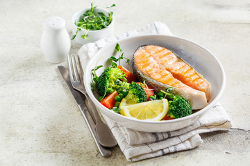 Grilled salmon steak with broccoli and tomatoes in white plate on gray background, copy space. Diet food concept.