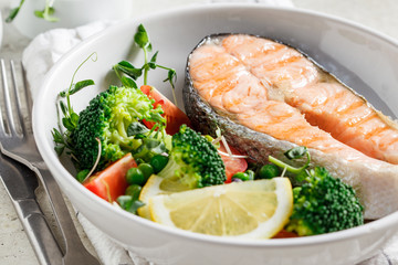 Grilled salmon steak with broccoli and tomatoes in white plate on gray background. Diet food concept.