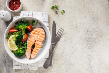 Grilled salmon steak with broccoli and tomatoes in a white plate on gray background, top view, copy space. Diet food concept.