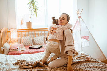 Fototapeta premium Beautiful young woman and her charming little daughter are hugging and smiling. Mother and her daughter child girl playing and hugging. Happy loving family.