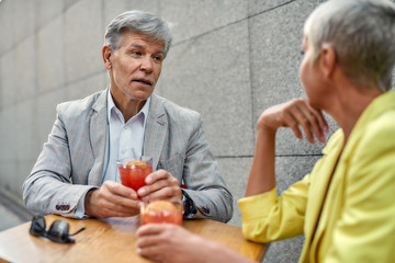 Handsome mature man in formal wear holding cocktail and discussing something with his female friend while sitting in cafe outdoors together