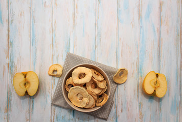 Dried apple chips in a bowl on a white wooden background and towel. Diet snack with copy space. Top view