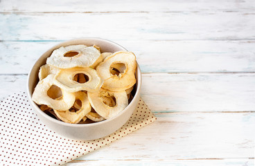 Dried apple chips in a bowl on a white wooden background and towel. Diet snack with copy space.