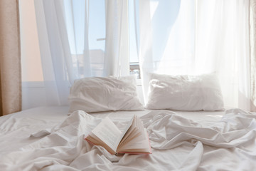 Book on bed with white linen in front of window with morning light through curtains