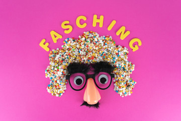 Funny face mask with big eyes and confetti hair, pink background with copy space and the German word for carnival in yellow letters