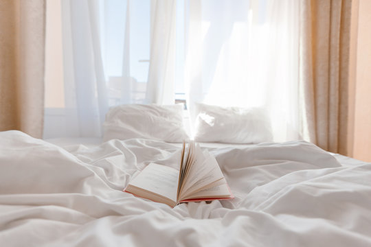 Book on bed with white linen in front of window with morning light through curtains