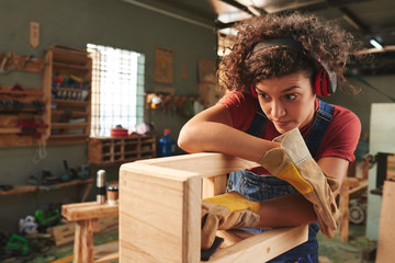Young curly woman in ear defenders and protective gloves checking quality of wooden stool after...