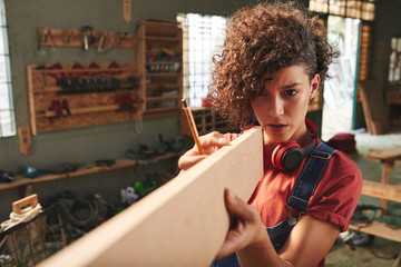 Young concentrated female carpenter with curly hair holding wooden plank and estimating its length...