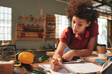 Young attractive female carpenter thoroughly checking measurements on blueprint before working with wood