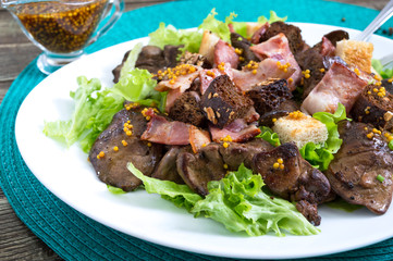 Warm healthy salad of chicken liver, rye croutons, smoked bacon, green salad and mustard sauce in a white plate on a wooden background. Close up