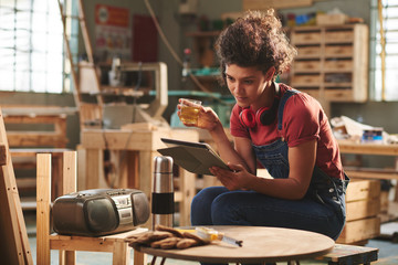 At carpentry workshop. Young pretty female carpenter in denim overall resting on wooden bench with...