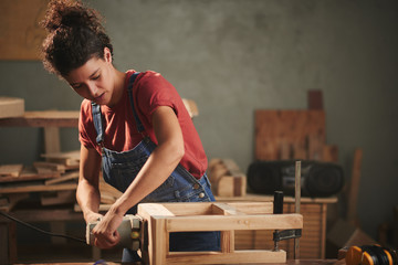 Young concentrated woman in denim overall smoothing just finished wooden stool with electrical belt...