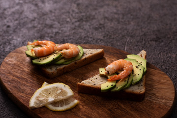 Wholegrain toast bread slices with guacamole, fried shrimp and fried bacon pieces on wooden board Selective Focus, Focus on the front of the shrimp on the first bread