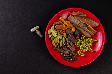 Top view of assorted whole spices, cinnamon, cardamom, pipali, cloves and two herbal capsules on a black background with copy space. Herbal medicines concept