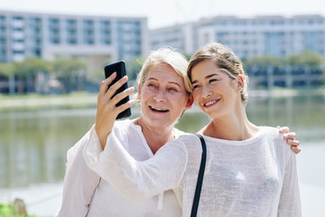 Pretty young woman talking selfie with her mature mother when they are walking outdoors on sunny day