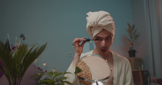 Young transgender applying mascara on eyelashes with opened mouth. Man trans in bath robe doing makeup and looking in mirror while getting ready for party. Room background.