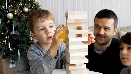Family is playing in board game with wooden tower together at home. Junior son is pulls out a brick while older brother and father is looking on it. - obrazy, fototapety, plakaty