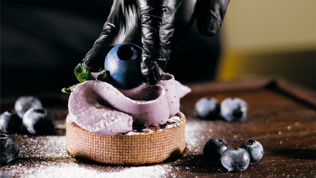 Pastry chef is puts a berry on top of cake, close-up. Professional baker is making blueberry cake in commercial bakery