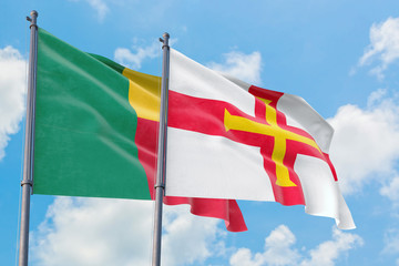 Guernsey and Benin flags waving in the wind against white cloudy blue sky together. Diplomacy concept, international relations.
