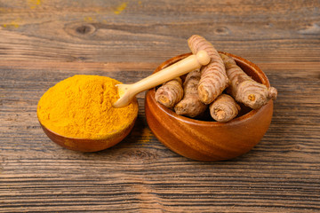 Fresh whole turmeric roots and dried curcuma powder in a wooden bowl in front of a wooden rustic background.