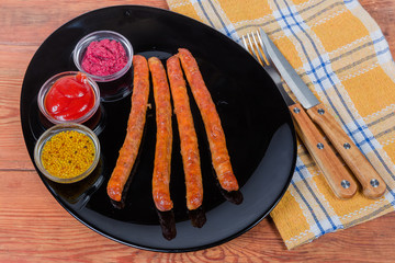 Baked pork sausages on black dish, various condiments, fork, knife