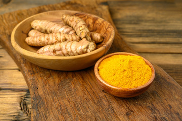 A wooden bowl with curcuma powder and a bowl with whole turmeric roots on a wooden cutting board.