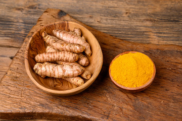 A wooden bowl with curcuma powder and a bowl with whole turmeric roots on a wooden cutting board.