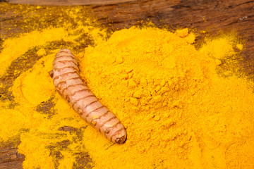 One fresh turmeric root an dried curcuma powder in front of a wooden background.