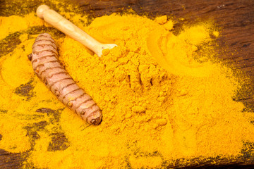 Dried curcuma powder with a wooden spoon and a whole fresh turmeric root in front of a wooden background.