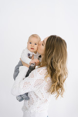 mother holds her five-month-old son in her arms against a light background and kisses him on the cheek