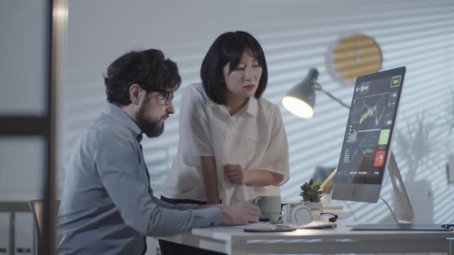 PAN of bearded businessman and Asian businesswoman looking at financial charts on computer screen and discussing data in dark office in evening
