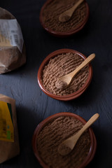 Fototapeta na wymiar Toasted carob flour, brown ceramic bowls, wooden spoons, all on a black background. Montuiri, Mallorca, Spain