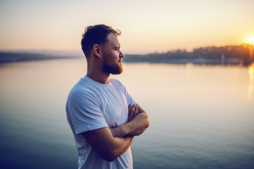 Handsome bearded blond caucasian man standing on cliff with arms crossed and looking at beautiful sunset. In background is river. - Powered by Adobe