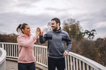 Young happy sporty heterosexual friends in sportswear standing on the bridge and giving high five...