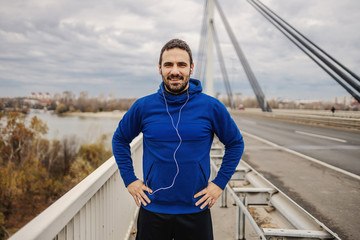 Attractive smiling young sportsman standing on the bridge and holding hands on hips. In ears are earphones. Outdoor fitness on cloudy weather concept. Urban exterior.