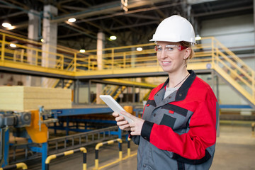 Portrait of cheerful young woman in protective goggles and hardhat checking data on tablet at construction plant