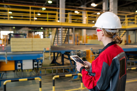 Rear view of busy woman in hardhat and safety goggles using tablet while controlling production line process at factory